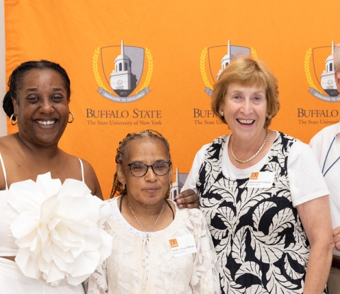 Group of five alumni smiling for the camera in front of an orange Buffalo State logo backdrop