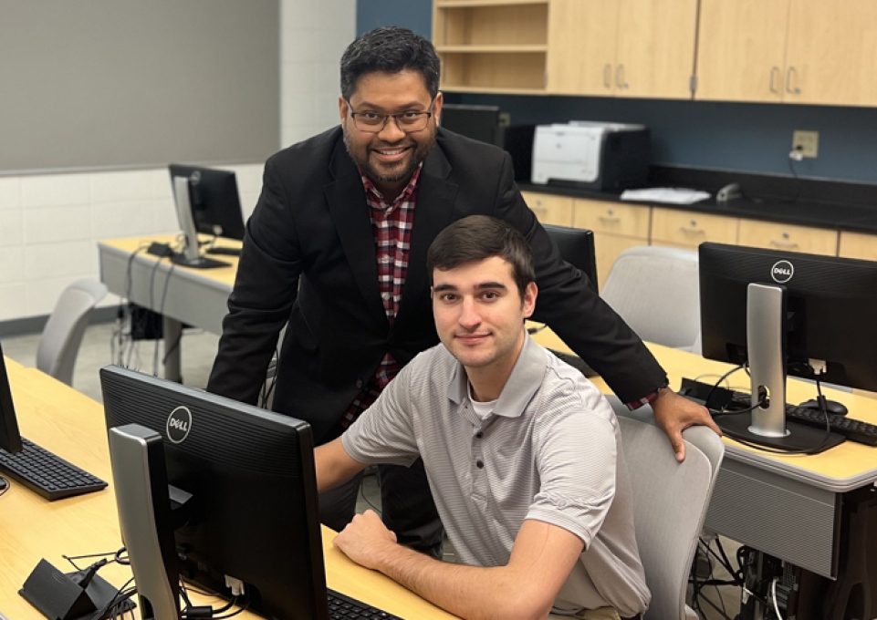 Saquib Ahmed stands behind Joseph Wikar, who is seated in front of a computer screen.